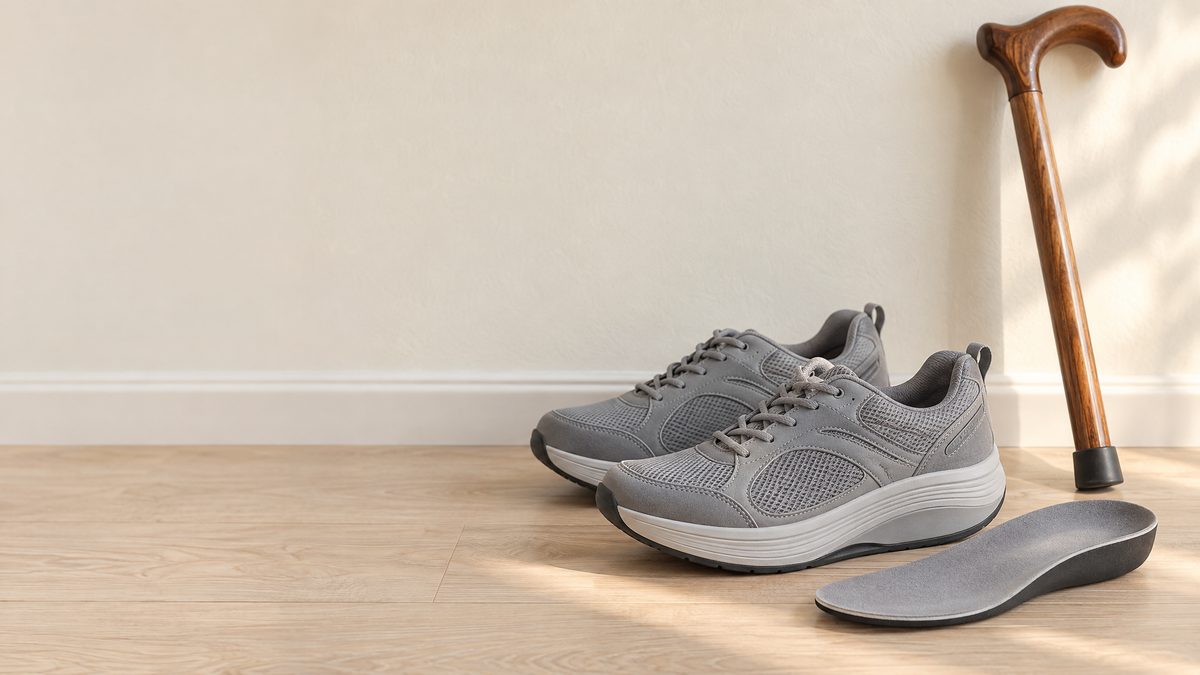 Gray walking shoes with rocker soles, a molded insole and a wooden cane against a pale wall